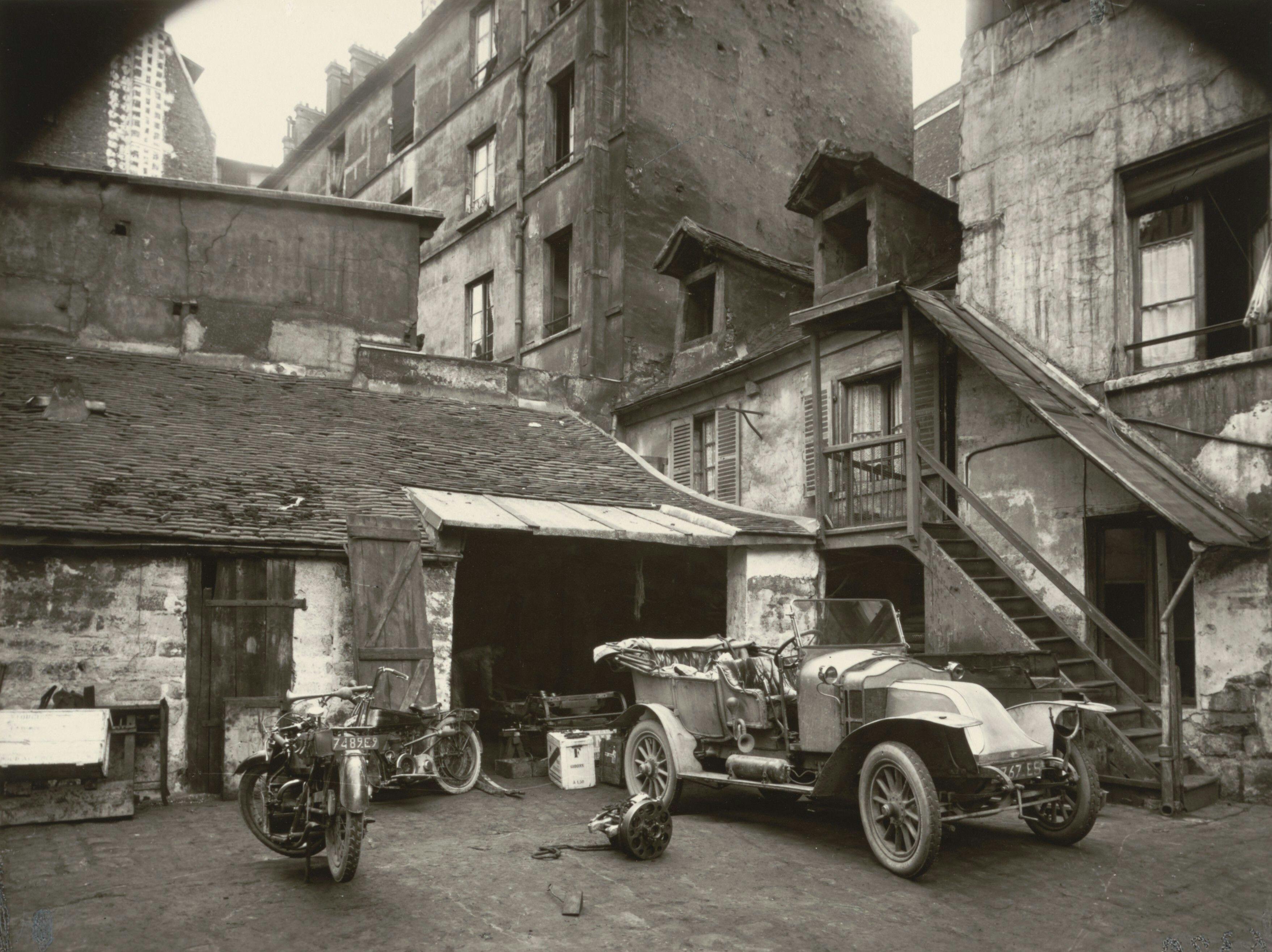 old photograph of a cluttered courtyard with a dilapidated building, vintage cars, motorcycles, and a wooden staircase, by Eugène Atget